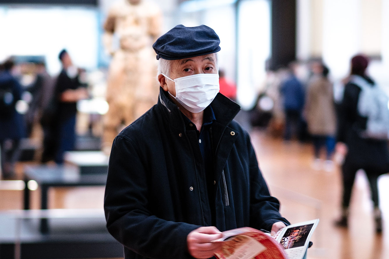 Older man with mask, holding a newspaper is sitting in a public hall with people in the background